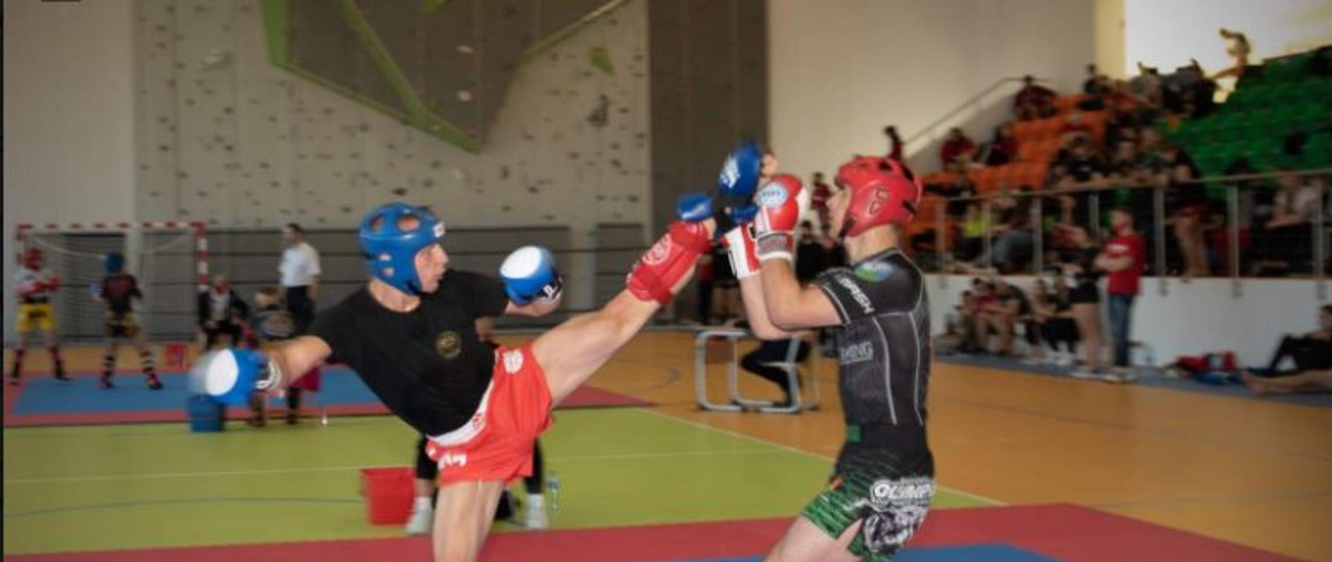 Zdjęcie przedstawia Adriana Durmę podczas walki na macie. Adrian uderza przeciwnika nogą, a ten próbuje zablokować uderzenie podniesionymi ręk0ma. W tle widoczne są trybuny i przyglądający się walce inni zawodnicy.
