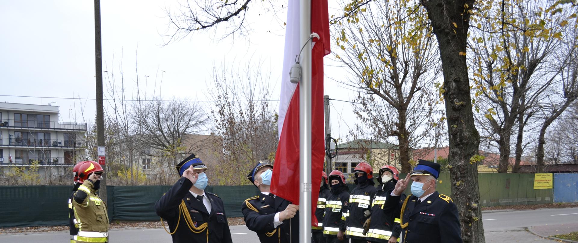 Przy maszcie flagowym stoi trzech strażaków w granatowych mundurach galowych, na głowach maja rogatywki, twarze zasłonięte maseczkami. Jeden z nich podnosi (wciąga) flagę państwową na maszt pozostali dwaj oddają honory fladze państwowej(salutują). Około dwa metry za masztem flagowym po lewej i prawej stronie stoi po czterech strażaków w ubraniach specjalnych w kolorach granatowym i piaskowym, którzy również oddają honory fladze państwowej. Na głowach mają czerwone hełmy, twarze zasłonięte kominiarkami. W tle widać ulicę, ogrodzenie, zarośla, drzewa oraz budynki. 