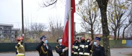Przy maszcie flagowym stoi trzech strażaków w granatowych mundurach galowych, na głowach maja rogatywki, twarze zasłonięte maseczkami. Jeden z nich podnosi (wciąga) flagę państwową na maszt pozostali dwaj oddają honory fladze państwowej(salutują). Około dwa metry za masztem flagowym po lewej i prawej stronie stoi po czterech strażaków w ubraniach specjalnych w kolorach granatowym i piaskowym, którzy również oddają honory fladze państwowej. Na głowach mają czerwone hełmy, twarze zasłonięte kominiarkami. W tle widać ulicę, ogrodzenie, zarośla, drzewa oraz budynki. 