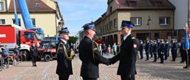 Na pierwszym planie trzej strażacy w czarnych mundurach galowych uczestniczą w gratulacjach; dwóch z nich podaje sobie ręce, podczas gdy trzeci stoi obok. W tle, po prawej stronie, ustawiony jest szereg strażaków w mundurach wyjściowych, a po lewej widoczne są pojazdy pożarnicze, w tym czerwony wóz bojowy oraz niebieski dźwig z uniesionym wysięgnikiem. Scenerię dopełniają zabytkowe kamienice oraz obserwatorzy w głębi placu.