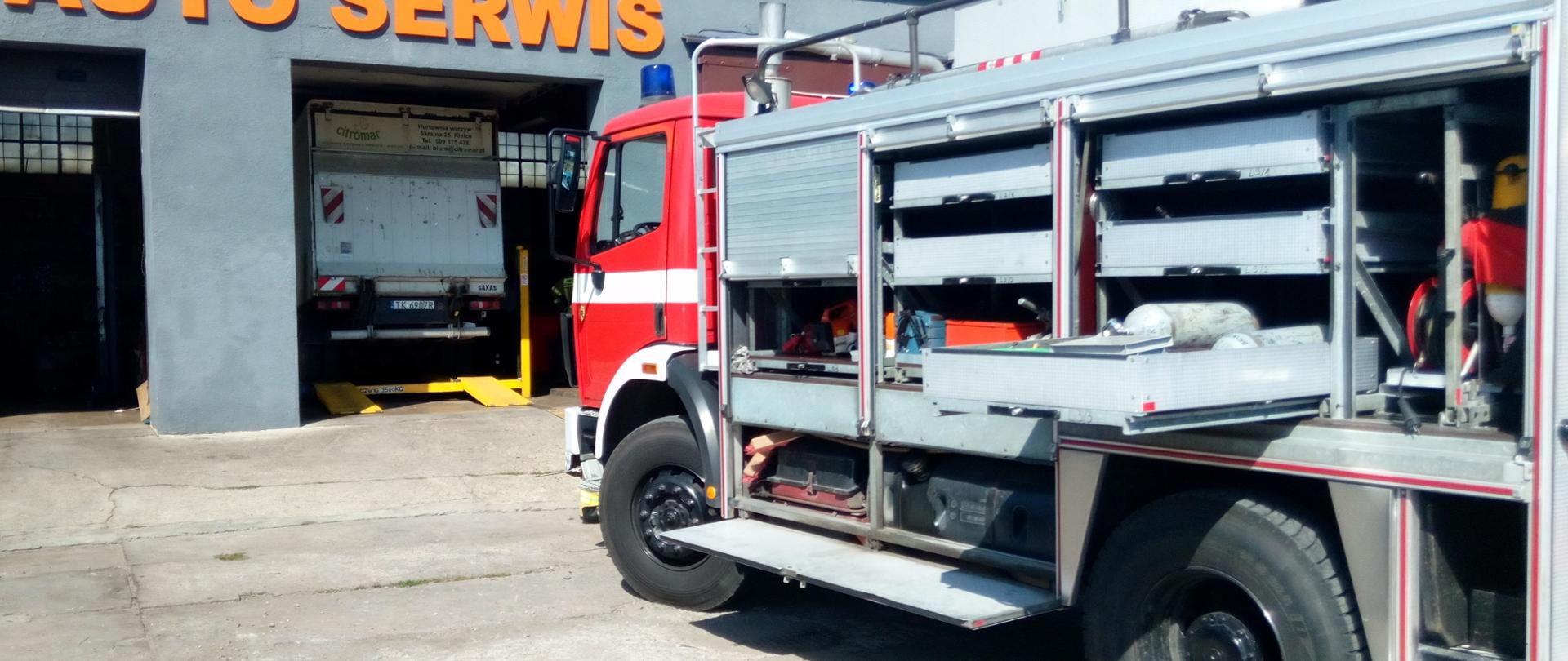 Na zdjęciu widzimy na pierwszym planie samochód pożarniczy podczas prowadzenia działań ratowniczych. W dalszej perspektywie widać budynek warsztatu samochodowego, a w otwartej bramie stoi samochód dostawczy, który przycisnął mężczyznę do ściany.