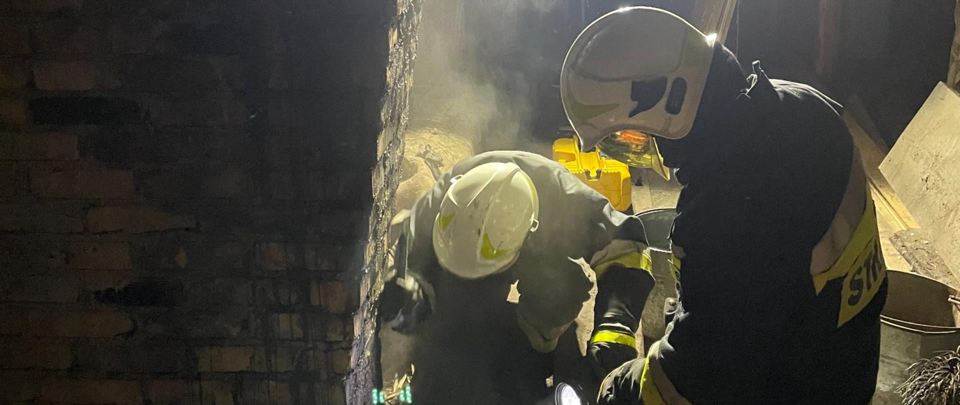 Okopcony komin. Przy kominie dwóch strażaków w pozycji klęczącej prowadzi prace rozbiórkowe stropu.