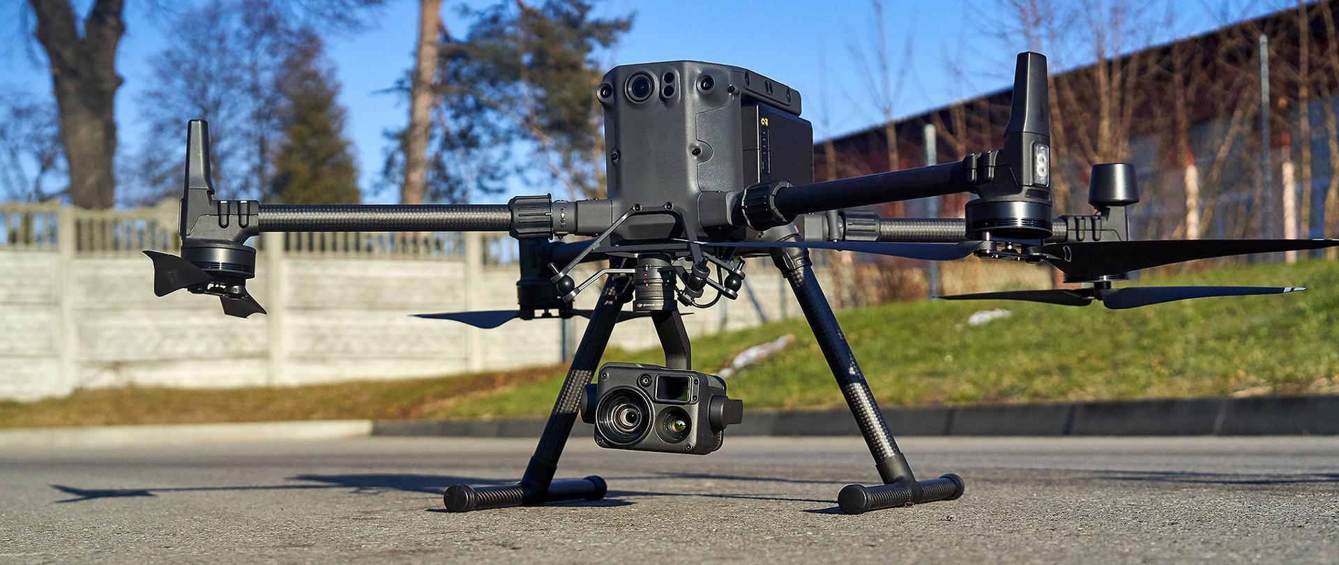 Na zdjęciu widoczny jest czterowirnikowy bezzałogowy statek powietrzny (dron). Do drona podczepiona jest prostokątna kamera z trzema obiektywami. Dron stoi na asfaltowej drodze. W środku kadru dobrze widoczny plastikowy garb, na którym umieszczone są czujniki w postaci niewielkich okręgów przypominających obiektywy.