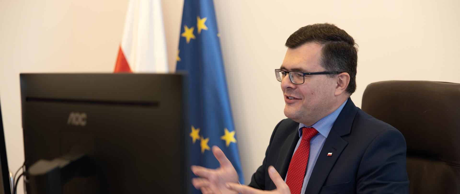 Minister Piotr Uściński w marynarce i niebieskiej koszuli z czerwonym krawatem siedzący za biurkiem przed monitorem na konferencji , gestykuluje rękami i uśmiecha się.