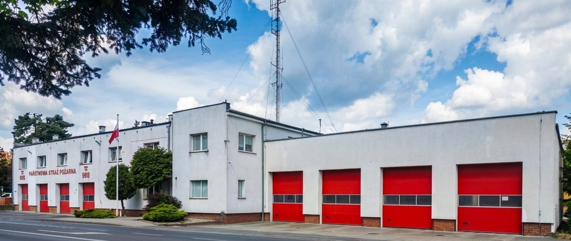 Budynek Komendy Powiatowej Państwowej Straży Pożarnej w Bolesławcu widziany od strony ulicy w słoneczny dzień. Biała, dwupiętrowa elewacja z napisem „PAŃSTWOWA STRAŻ POŻARNA” oraz numerem alarmowym 998, obok której wznosi się maszt z polską flagą. Po prawej i po lewej stronie widoczne są po cztery duże, jaskrawoczerwone bramy garażowe dla wozów strażackich.