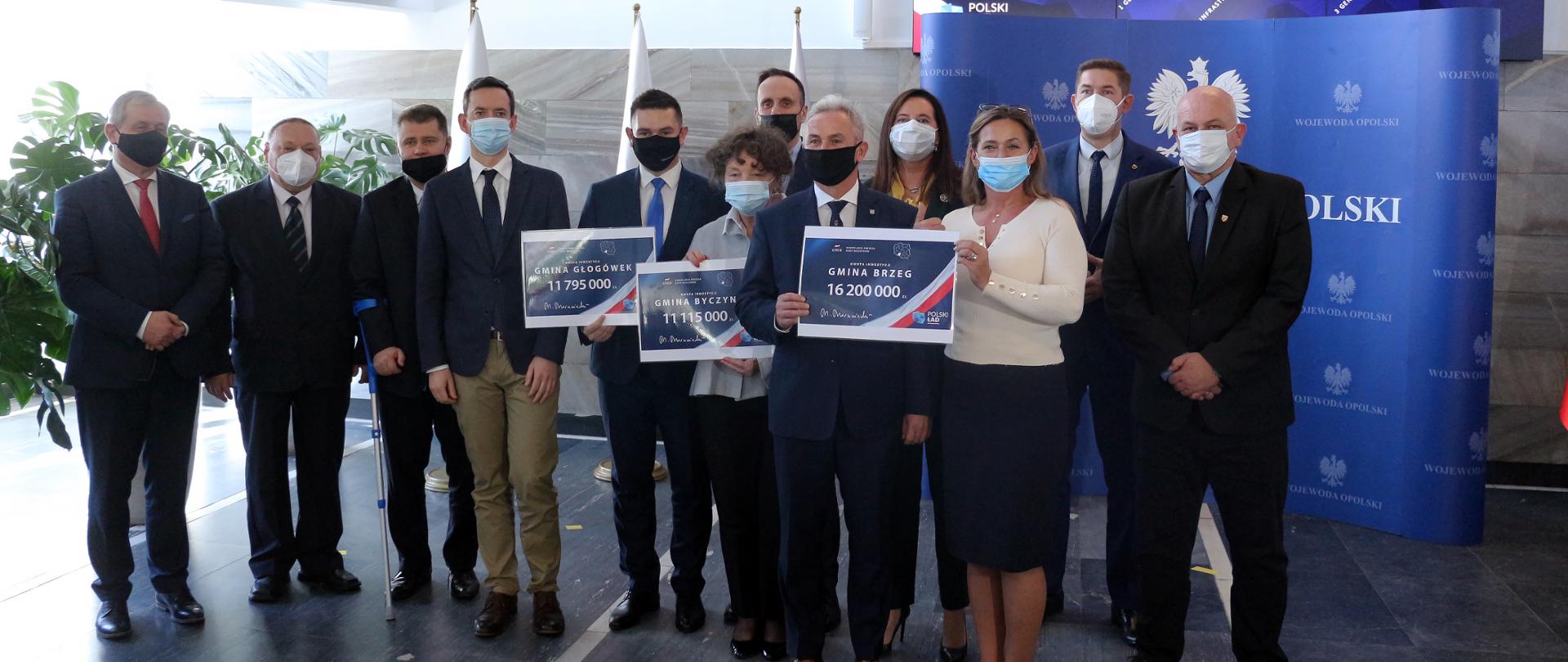 Na zdjęciu uczestnicy konferencji prasowej, która w dniu 25 października odbyła się w urzędzie wojewódzkim w Opolu. Zdjęcie przedstawia 13 osób stojących obok siebie, pozujących do wspólnej fotografii.. Od lewej stoi wojewoda opolski, następnie przedstawiciele kilku opolskich samorządów, posłowie. W środku kadru wiceminister obrony narodowej Marcin Ociepa. W tle widać granatową ściankę z napisem Wojewoda Opolski oraz duży monitor, na którym wyświetlana jest prezentacja. 