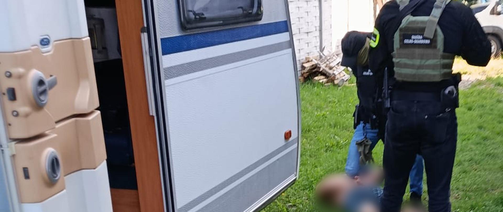 Funkcjonariusze Służby Celno-Skarbowej prowadzą czynności na posesji obok otwartej przyczepy kempingowej; na trawie widoczne są zamazane elementy obrazu.