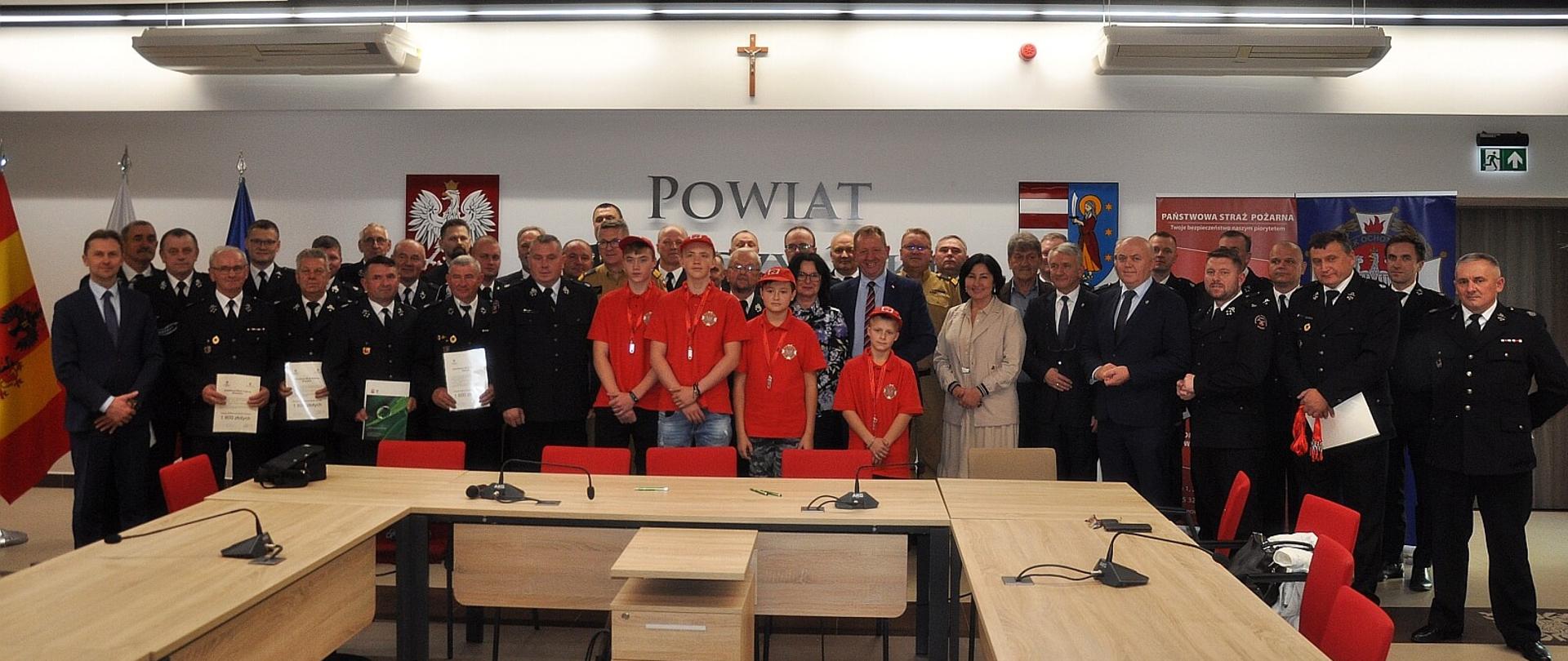uczestnicy uroczystości młodzież strażacy politycy i samorządowcy pozują do zdjęcia w sali, przez zgromadzonymi sa stoły za ściana z napisem "powiat"