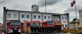 ZDJĘCIE PRZEDSTAWIA STRAŻAKÓW W UMUNDUROWANIUE PODCZAS POZYCJI ZASADNICZEJ NA TLE KOMNEDY I AUT POŻARNICZYCH PODCZAS ODDAWANIA HOŁDU