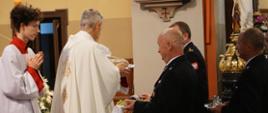 Zdjęcie przedstawia Strażaków, którzy przynoszą dary do ołtarza. Dary odbiera Ksiądz Kapelan. Dary składane w ofierze widoczne na zdjęciu to chleb i winogrono. Dary składa Komendant Powiatowy PSP w Łańcucie oraz druh Adam Reizer prezes zarządu powiatowego ZOSP.