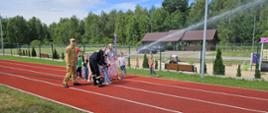 Na tartanowej bieżni sportowej grupa dzieci w asyście dwóch strażaków PSP i Pani nauczycielki leją wodę z węża strażackiego