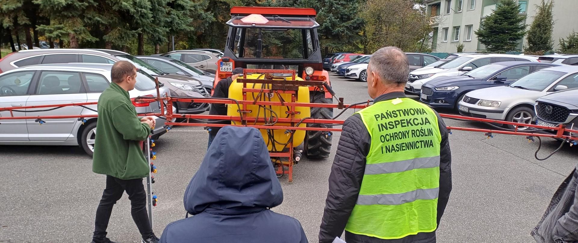 
Kilka osób stojąca na zewnątrz. Przed nimi stoi traktor. Mężczyzna po prawej ma ubraną odblaskową kamizelkę.
