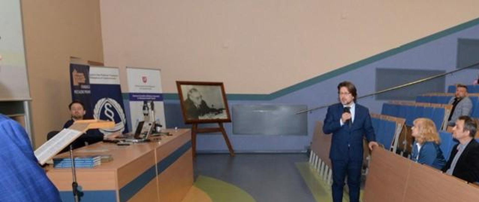 Dr. Jarosław Reterski, ubrany w niebieski garnitur, włosy ściągnięte do tyłu, dłuższe, podczas swojego wystąpienia konferencja "EKO-JURA – Nauka Przedsiębiorcom"