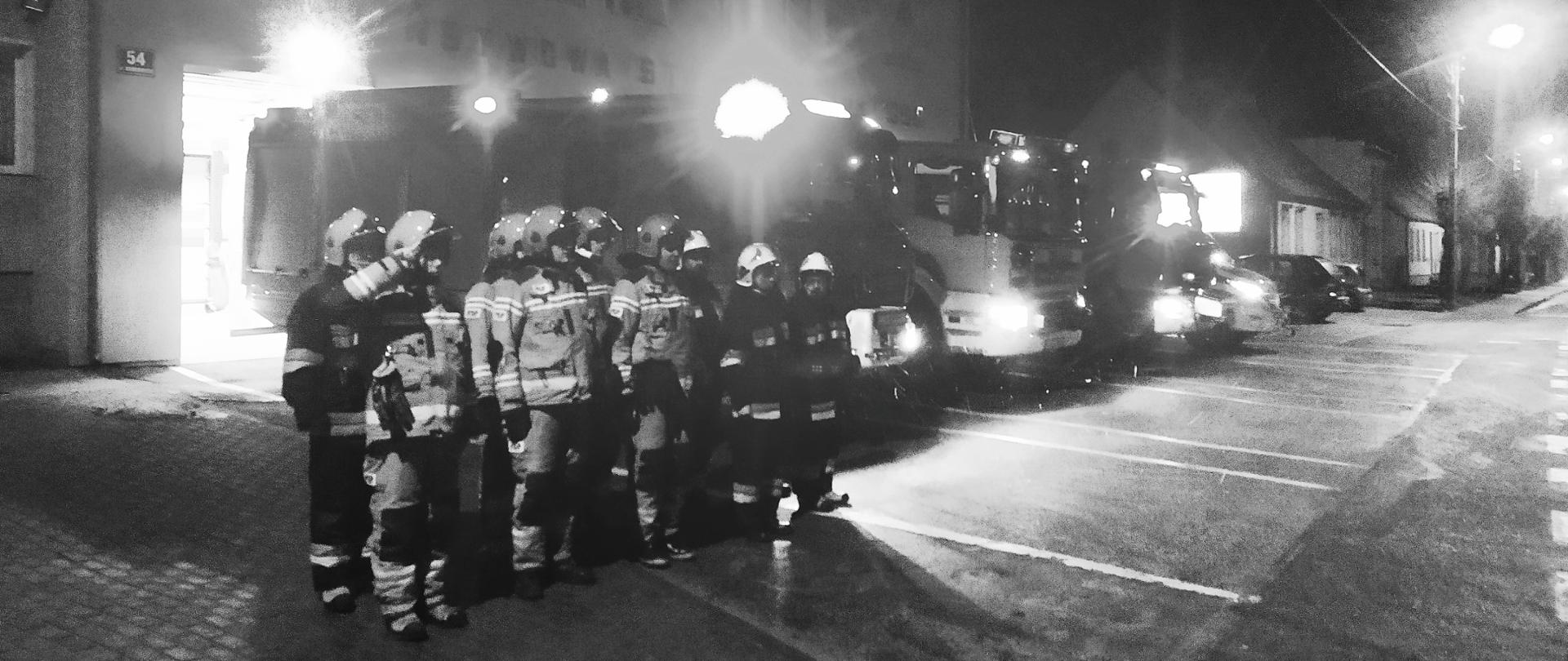 Zdjęcie wykonano wieczorem przed siedzibą Komendy Powiatowej Państwowej Straży Pożarnej w Sępólnie Krajeńskim. Strażacy Państwowej oraz Ochotniczej Straży Pożarnej stoją w dwuszeregu ubrani w ubrania specjalne oraz hełmy. Za nimi stoją samochody ratowniczo- gaśnicze, w tle budynek Komendy Powiatowej. 