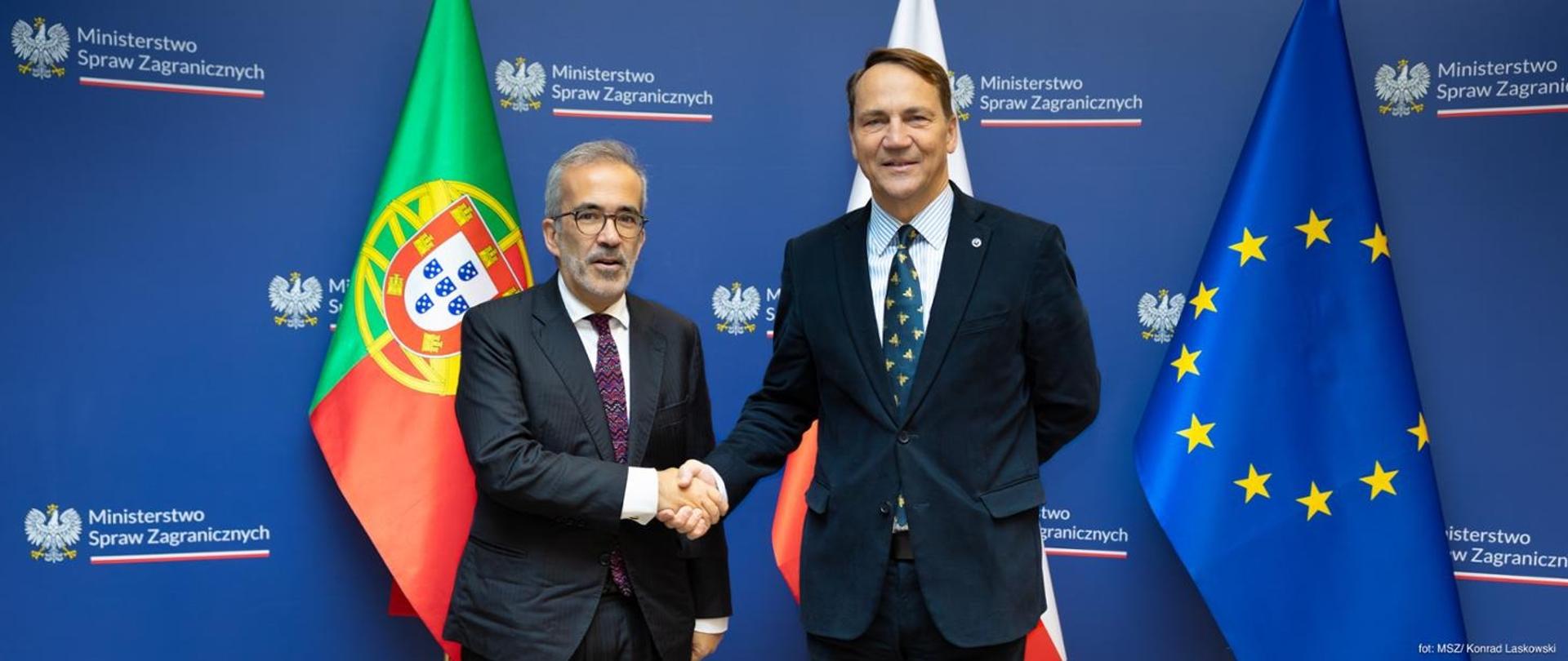 Primeiro-Ministro Adjunto da Polónia, Ministro dos Negócios Estrangeiros Radosław Sikorski e o Ministro de Estado e dos Negócios Estrangeiros de Portugal, Paulo Rangel, estão lado a lado diante de bandeiras: à esquerda a bandeira de Portugal, à direita a bandeira da União Europeia e parte da bandeira da Polónia. Ambos sorriem para a câmara enquanto apertam as mãos num gesto de saudação. Ao fundo vê-se a decoração do espaço e o logótipo do Ministério dos Negócios Estrangeiros.