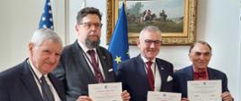 Na sali stoi w rzędzie czterech mężczyzn trzymających kartki z napisem Certificate of Appreciation, drugi od lewej wiceminister Szeptycki, za nimi na białej ścianie wisi obraz w złoconych ramach.