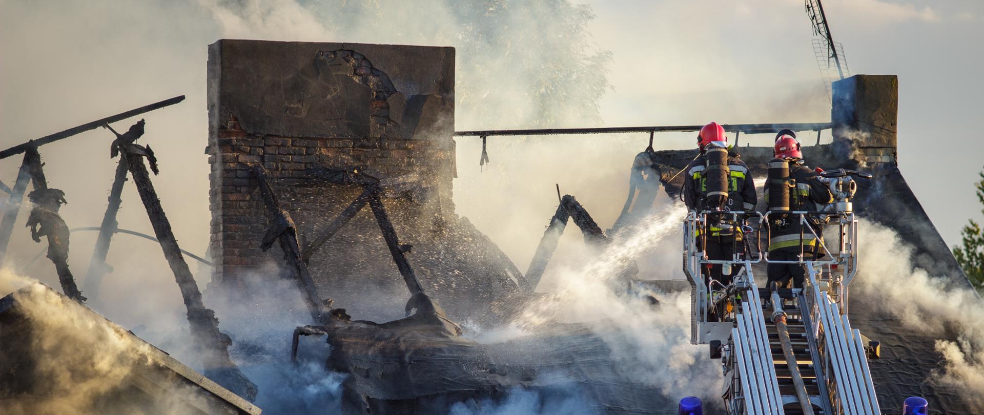 Dwóch strażaków w strojach bojowych na wysuwanej drabinie gasi resztki ognia na dachu domu. Cały dach się zapadł biały dym snuje się poniżej czarnych osmolonych belek dachowych i dwóch murowanych kominów.