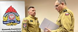 Dwóch mężczyzn w mundurach Państwowej Straży Pożarnej stoi w biurze. Po prawej stronie jeden z nich komendant powiatowy starszy kapitan Mirosław Śledź trzyma w rękach dokument, decyzję o nadaniu nagrody jubileuszowej i czyta ją, zwracając się do drugiego strażaka, który stoi naprzeciwko niego w postawie zasadniczej. W tle po lewej znajduje się roll-up z logo PSP oraz informacjami kontaktowymi Komendy Powiatowej Państwowej Straży Pożarnej w Szczecinku. Po prawej stronie, przy ścianie, stoi niewielki stojak z herbami lokalnych jednostek. Scena jest podczas wręczenia nagrody jubileuszowej.