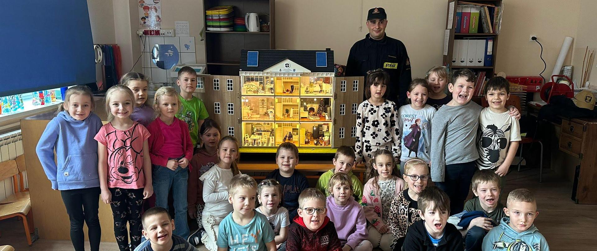 Zdjęcie grupowe strażaka z symulatorem zagrożeń oraz dziećmi uczestniczącymi w pogadance. Zdjęcie wykonane w klasie lekcyjnej. Uśmiechnięte dzieci pozują ze strażakiem upamiętniając wizytę przedstawiciela komendy w placówce dydaktycznej.