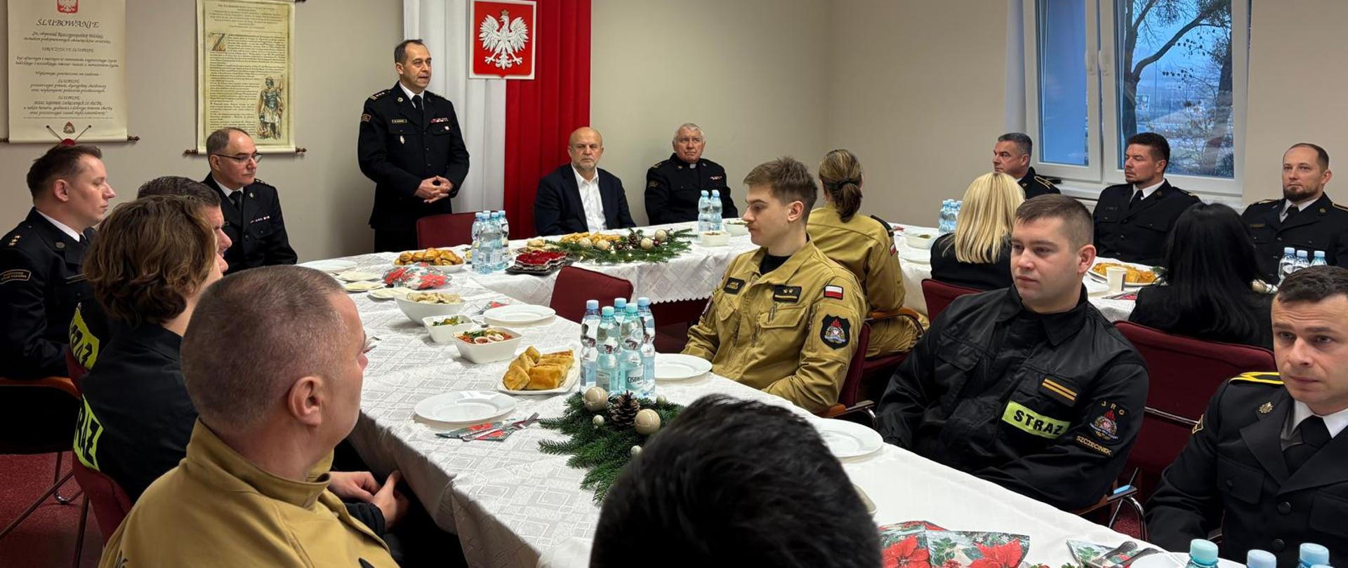 Uczestnicy spotkania wigilijnego Państwowej Straży Pożarnej zgromadzeni przy wspólnym stole w sali konferencyjnej jednostki. W uroczystej atmosferze, przy tradycyjnych potrawach, strażacy oraz kadra dowódcza wspólnie celebrowali przedświąteczny czas, dzieląc się życzeniami i refleksją nad mijającym rokiem służby. Ubrani w mundury wyjściowe koloru granatowego oraz piaskowe.