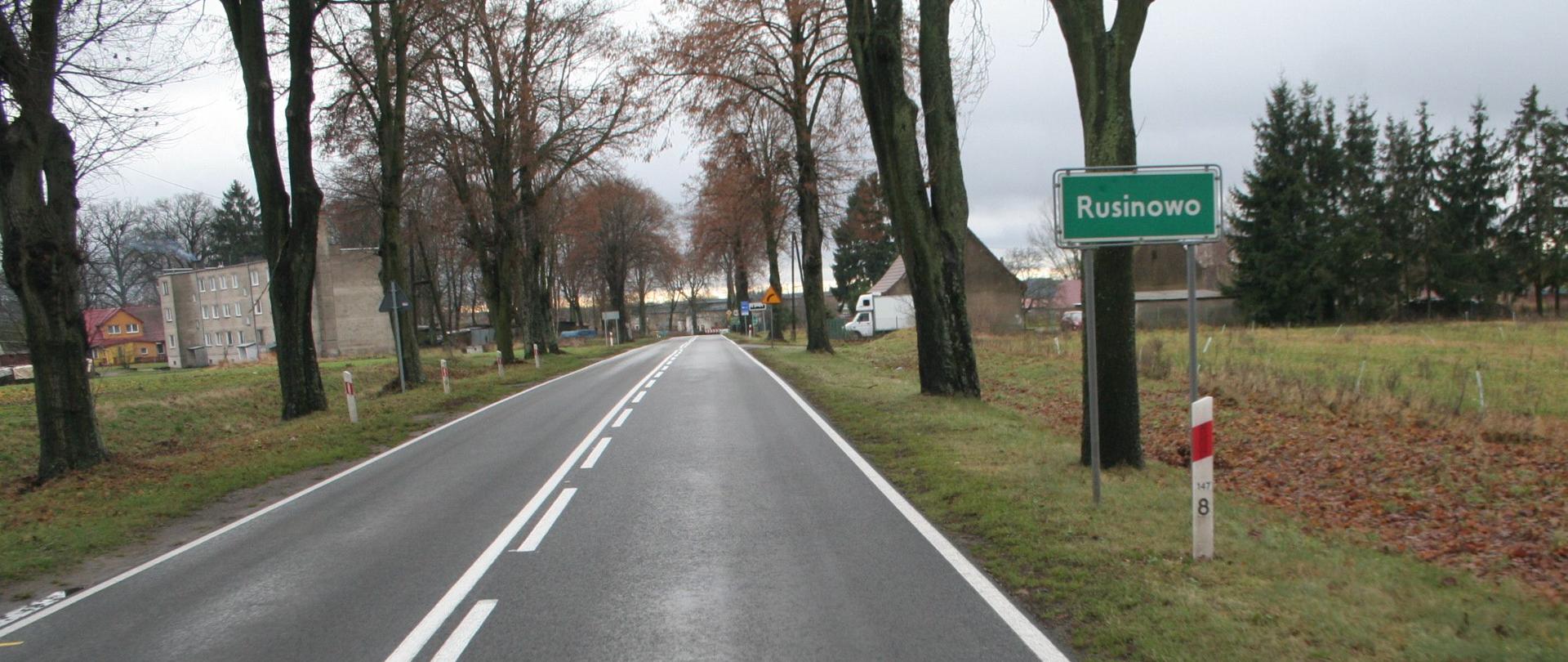 Wjazd drogą krajową nr 22 do miejscowości Rusinowo. Przy drodze po obu stronach rosną wysokie liściaste drzewa. W tle zabudowania Rusinowa.