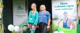 Przy stoisku informacyjno-promocyjnym Kasy pozują do zdjęcia dwie kobiety. Za nimi znajduje się stół z materiałami promocyjnymi Kasy oraz dekoracje (roll-upy, kwiaty, balony).