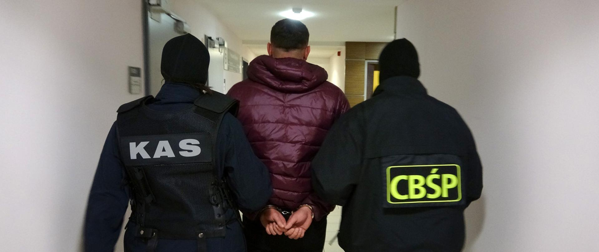 Funkcjonariusze KAS i CBŚP prowadzą zatrzymanego mężczyznę w kajdankach.