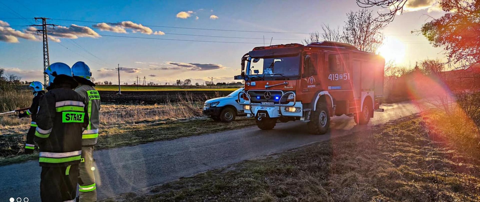 Na zdjęciu wykonanym przy zachodzie słońca, po prawej części fotografii znajduje się wóz strażacki. Po lewej stronie stoi trzech strażaków. W oddali jest znajduje się pole oraz linie energetyczne.
