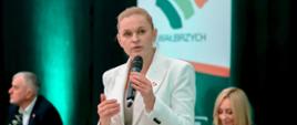 Minister Nowacka stoi, w ręku trzyma mikrofon i mówi. W tle kobieta i mężczyzna siedzą przy stole i słuchają.