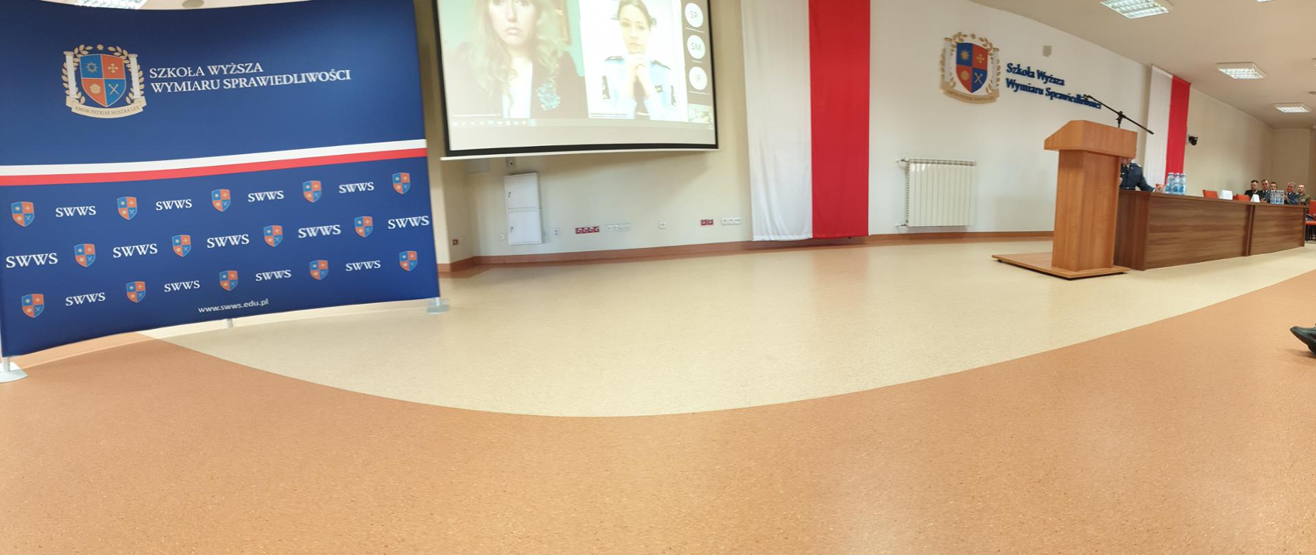 Sala sztucznie oświetlona. Widać flagę państwa, herb szkoły oraz nazwę. Drewniana mównica i stół. Po prawej uczestnicy konferencji siedzą na krzesłach. 