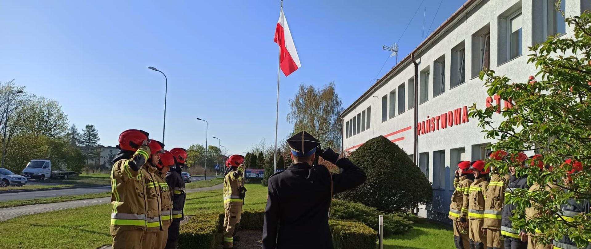 Zdjęcie przedstawia uroczystą zmianę służby przed budynkiem Komendy Powiatowej Państwowej Straży Pożarnej w Świdwinie. Trzech strażaków pod masztem flagowym oddaje honory do Flagi Państwowej. Przed asystą honorową znajdują się w szeregu równolegle do siebie zwrócone zmiany. Na środku znajduję się Komendant powiatowy w mundurze galowym.