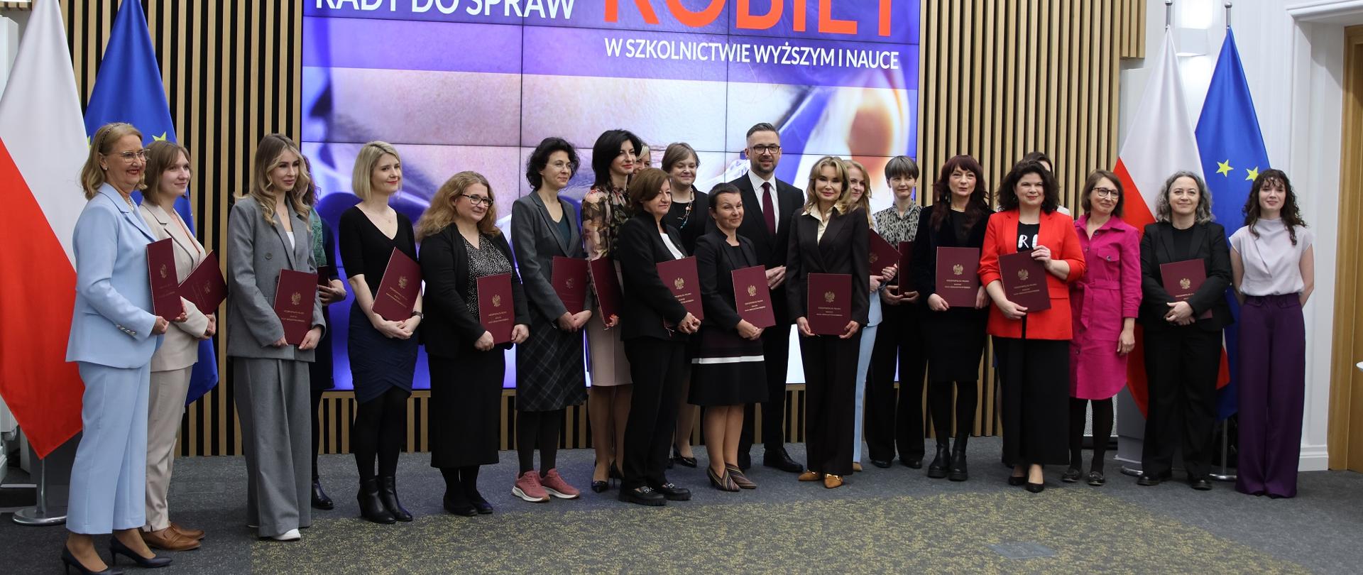 Zdjęcie zbiorowe, pod ścianą z pionowych drewnianych listewek z wbudowanym wielkim ekranem z napisem Powołanie Rady ds. Kobiet w Szkolnictwie Wyższym i Nauce stoi grupa kilkunastu kobiet trzymających czerwone teczki, pośrodku grupy minister Kulasek i wiceminister Zioło-Pużuk.