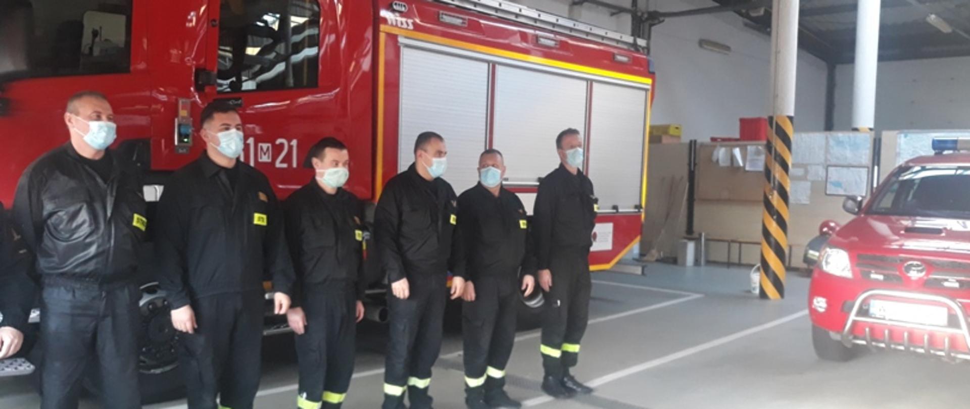 Zdjęcie przedstawia uroczystość wręczenia awansu strażakowi PSP. W jednym szeregu na tle samochodu gaśniczego w garażu Jednostki Ratowniczo Gaśniczej stoi pododdział ubrany w ubrania koszarowe.