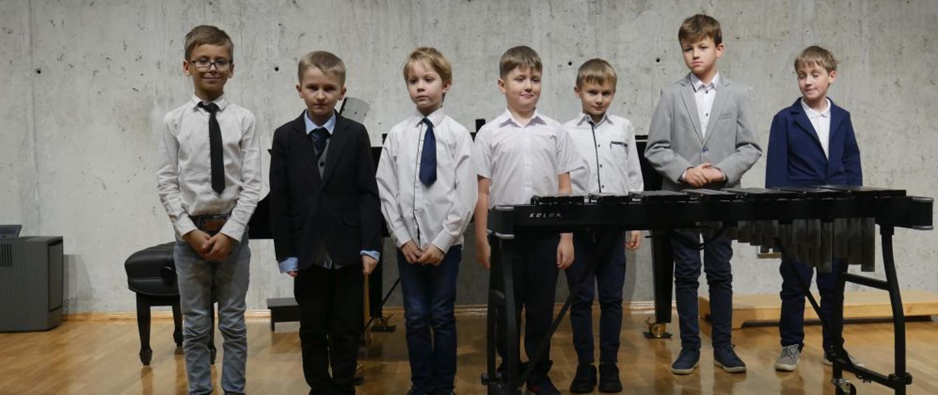 Zdjęcie przedstawia grupę elegancko ubranych chłopców stojących na scenie sali koncertowej, w tle fortepian, na pierwszym planie po prawej stronie przed chłopcami stoi ksylofon