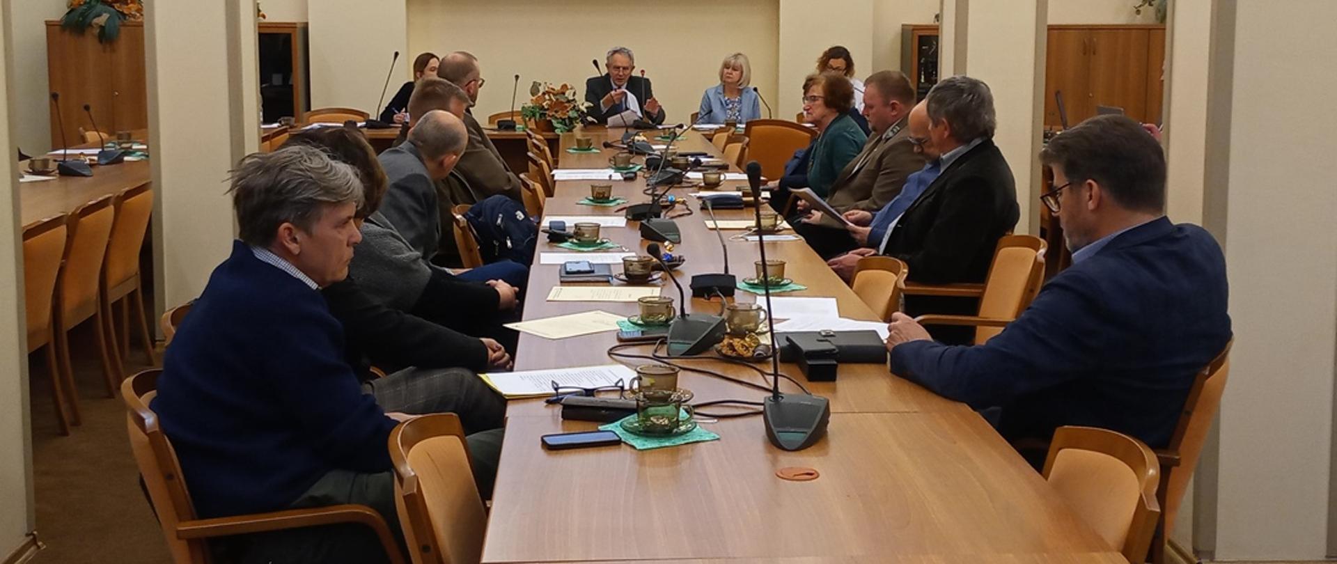 Duży stół, na którym stoją mikrofony oraz leżą dokumenty, wokół niego siedzą ludzie i dyskutują. 