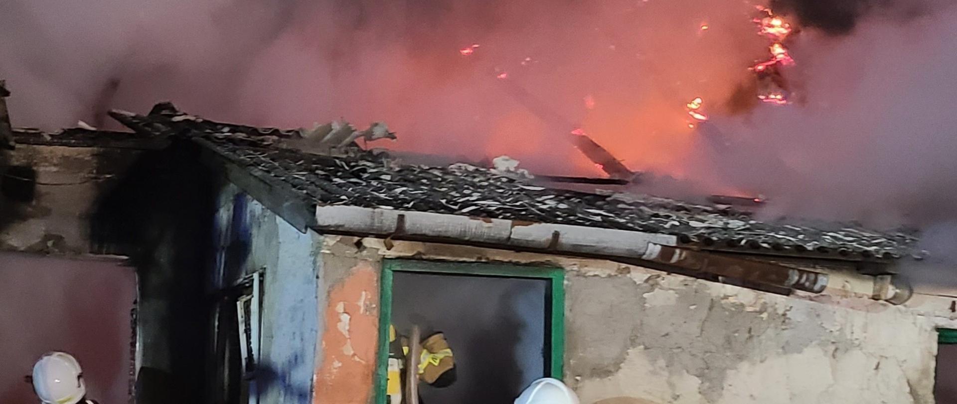 Zdjęcie przedstawia budynek, w którym doszło do pożaru. Nad dachem widać unoszący się dym i płomienie.