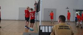 Na pierwszym planie widzimy funkcjonariusza PSP, który liczy punktu w meczu siatkówki. Dalej widać siatkę oraz zawodników trzymających ręce w górze. na tyle planu widoczny jest sędzia. 