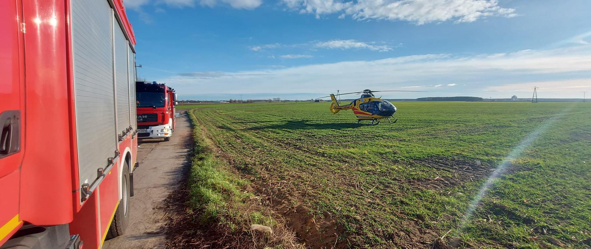 Śmigłowiec LPR stoi na polu obok na drodze stoją dwa samochody strażackie.