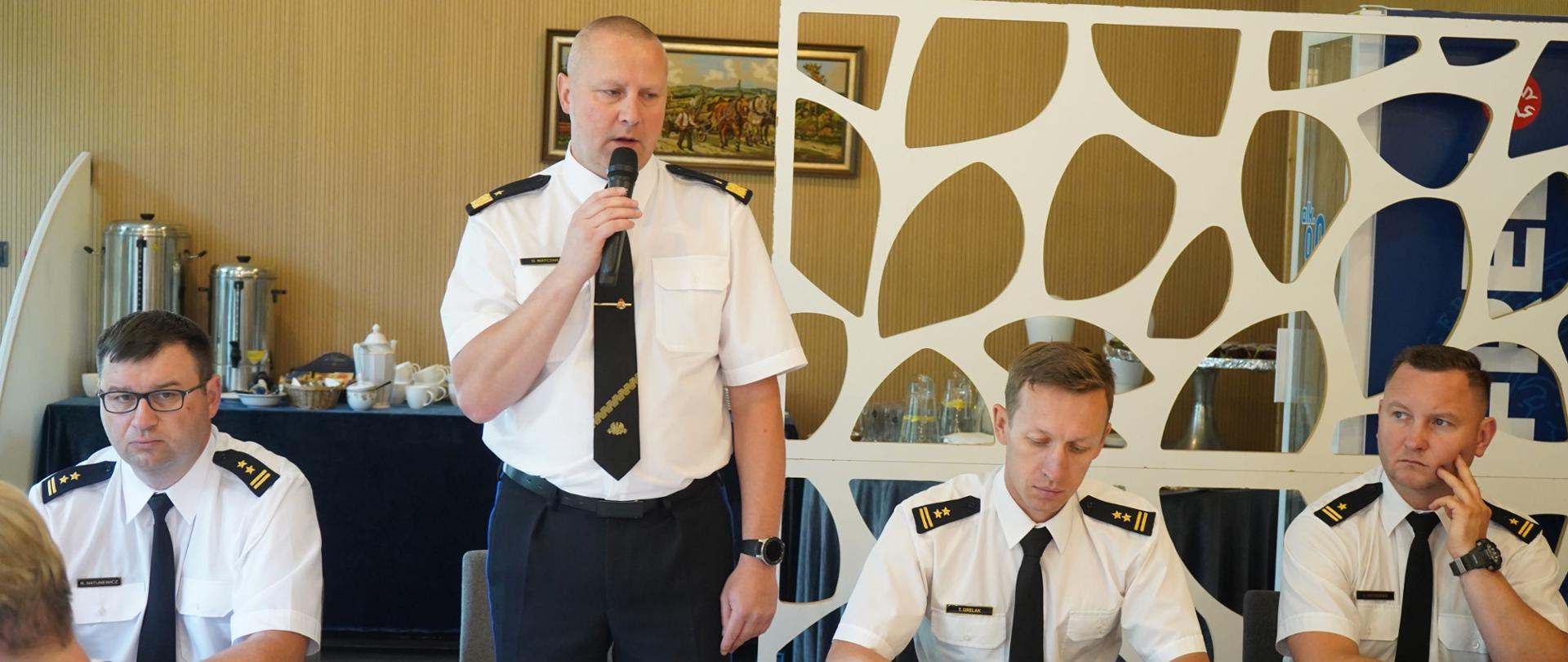jeden strażak stoi z mikrofonem przed stołem, przy tym stole siedzi jeszcze trzech strażaków, w tle jasnobrązowa ściana i obraz
