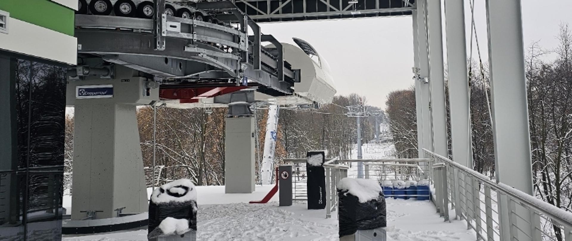 Stacja kolejki linowej z kabiną i ośnieżonym otoczeniem, w tle drzewa.