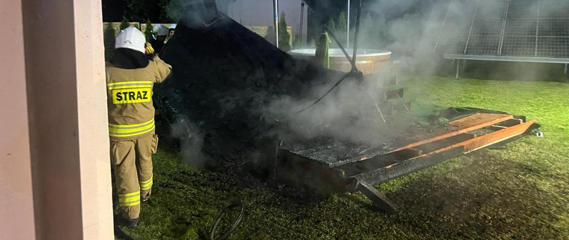 Zdjęcie przedstawia drewniany spalony budynek sauny, z którego wydobywa się biały dym. Koło budynku widać kilku strażaków w umundurowaniu specjalnym, którzy prowadzą działania gaśnicze. Zdjęcie zrobione w porze nocnej.