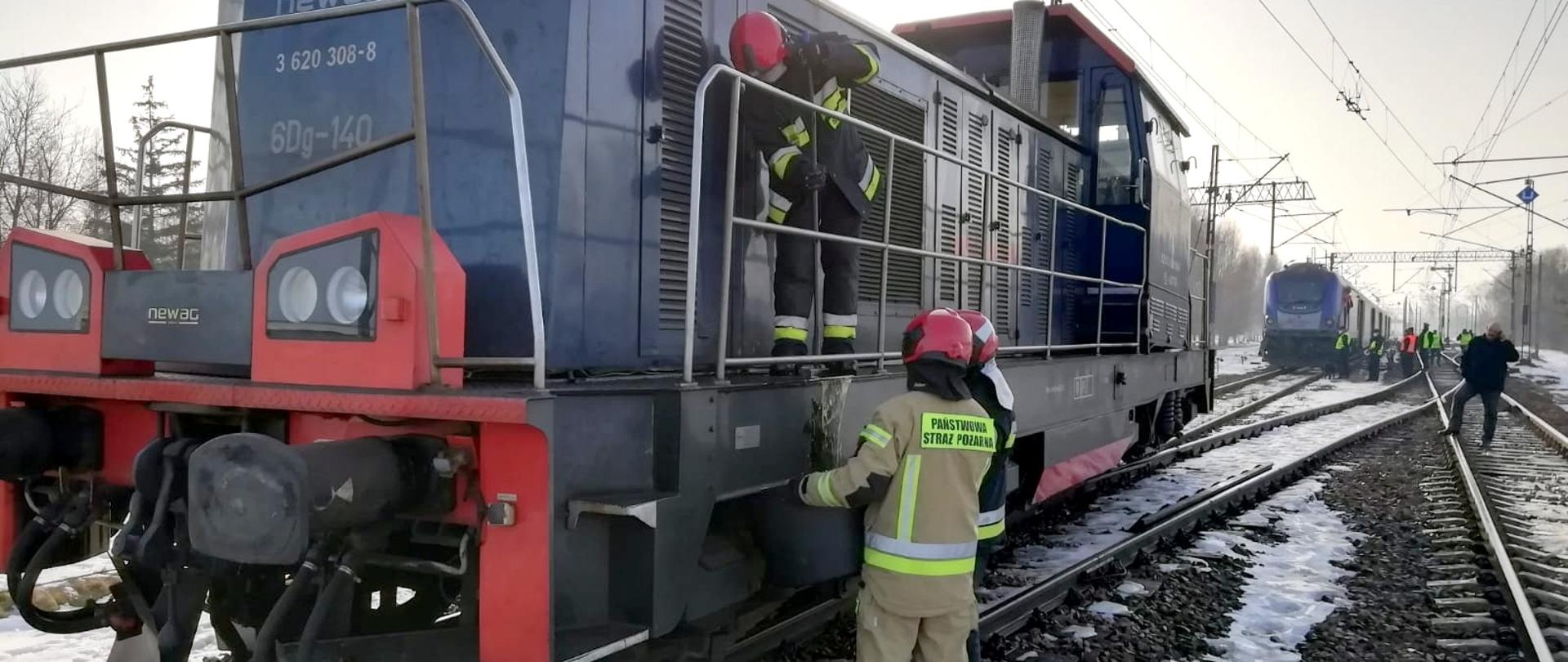 Na zdjęciu lokomotywa spalinowa. Obok lokomotywy oraz na pojeździe strażacy usuwają wyciekający olej napędowy. W tle, za lokomotywą stoi pociąg pasażerski.
