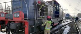 Na zdjęciu lokomotywa spalinowa. Obok lokomotywy oraz na pojeździe strażacy usuwają wyciekający olej napędowy. W tle, za lokomotywą stoi pociąg pasażerski.