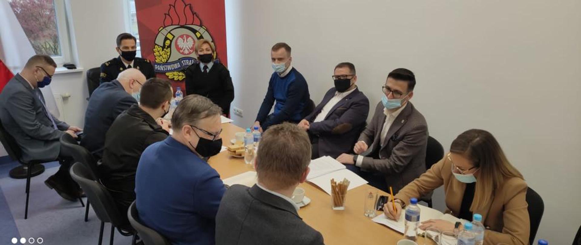 Na zdjęciu 11 osób siedzi przy wspólnym stole. Uczestnicy rozmawiają i notują. W tle znak państwowej straży pożarnej