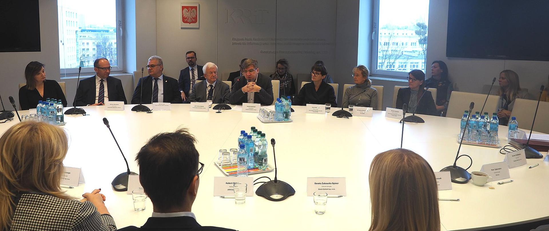  Zdjęcie zbiorowe. Członkowie KRRiT oraz przedstawiciele nadawców zasiadający przy okrągłym stole. W tle ściana z logo KRRiT