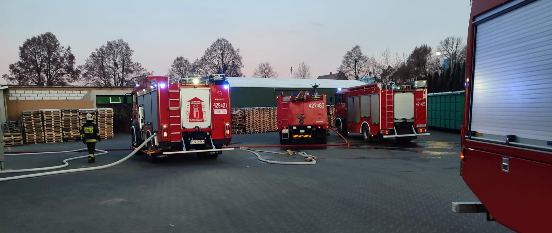 Na zdjęciach widać tyły trzech samochodów pożarniczych stojące na placu zakładu produkcyjnego. Z pojazdów podawana jest wężami pożarniczymi woda