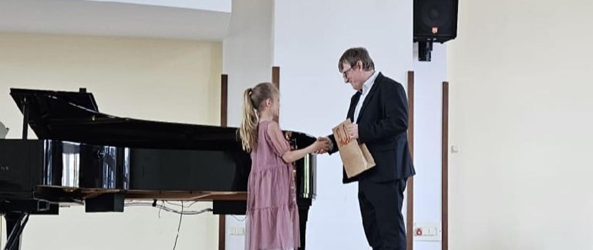 Zdjęcie kolorowe. Dziewczynka we fioletowej sukience przyjmuje na scenie gratulacje i nagrodę od mężczyzny w garniturze. W tle fortepian i głośnik