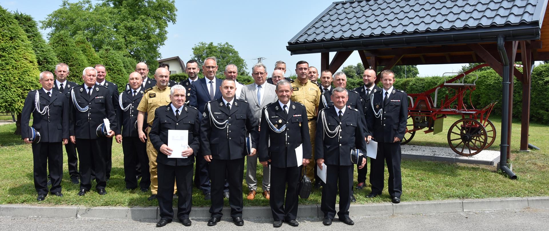 Zdjęcie grupowe uczestników spotkania. Wszyscy uczestnicy ubrani w stroje oficjalne, natomiast strażacy w umundurowanie wyjściowe. Po prawej stronie widoczna zabytkowa bryczka strażacka.