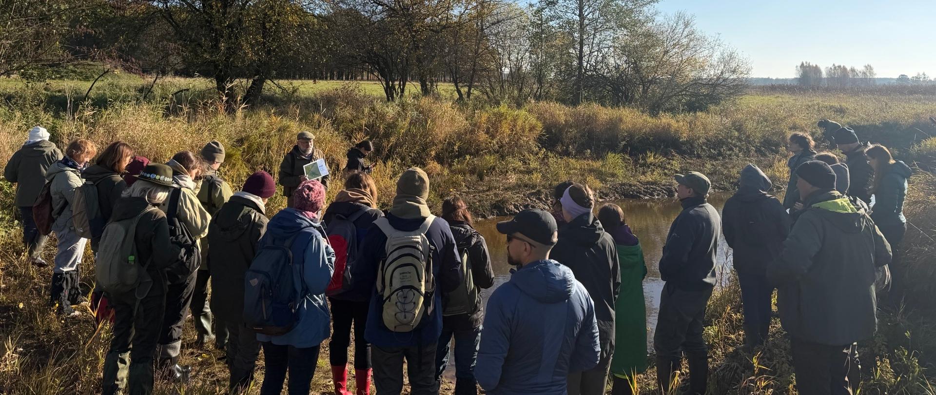 Zdjęcie przedstawia grupę uczestników terenowych warsztatów zgromadzonych nad niewielkim ciekiem wodnym. Osoby stoją na brzegu wśród naturalnej roślinności i słuchają prowadzącego. W tle widoczne są drzewa w jesiennej scenerii, a słoneczna pogoda podkreśla walory przyrodnicze odwiedzanego obszaru.

