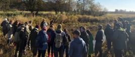 Zdjęcie przedstawia grupę uczestników terenowych warsztatów zgromadzonych nad niewielkim ciekiem wodnym. Osoby stoją na brzegu wśród naturalnej roślinności i słuchają prowadzącego. W tle widoczne są drzewa w jesiennej scenerii, a słoneczna pogoda podkreśla walory przyrodnicze odwiedzanego obszaru.
